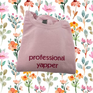 May include: Pink sweatshirt with the text "professional yapper" embroidered in maroon.