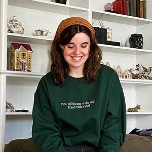 May include: A dark green crewneck sweatshirt featuring white embroidered text on the chest that reads, you tellin me a shrimp fried this rice? The garment has long sleeves and a ribbed neckline, providing a simple and functional design.