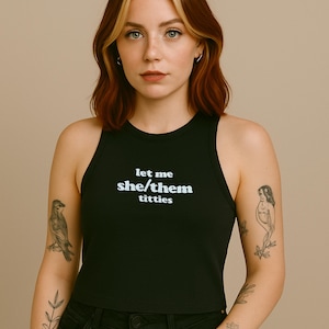 Op de afbeelding: Zwarte tanktop met de witte tekst "let me she/them titties". De top heeft een ronde halslijn en is mouwloos. De persoon op de foto draagt een zwarte jeans.