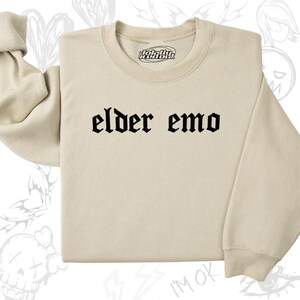 Può includere: Una felpa beige con la scritta "elder emo" stampata in caratteri gotici neri.