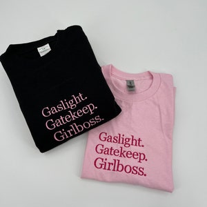 Puede incluir: Dos camisetas con el texto "Gaslight. Gatekeep. Girlboss." impreso en ellas. Una camiseta es negra con texto rosa y la otra es rosa con texto rosa oscuro.