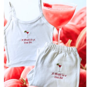 Könnte beinhalten: Weißes Pyjama-Set mit Cocktailglas-Stickerei und dem Text "A Bride in a tini bit". Das Set beinhaltet ein Tanktop und eine Hose. Ein rosa Cocktail mit Zuckerrand ist im Hintergrund.