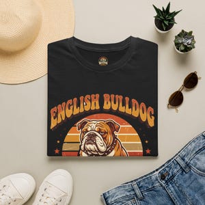 Könnte beinhalten: Ein schwarzes T-Shirt mit einer Grafik eines Englischen Bulldoggen, zentriert über einem retro-gestalteten Sonnenuntergangsbogen. Der Text "ENGLISH BULLDOG" ist in einer fetten, geschwungenen Schrift über dem Hundemotiv aufgedruckt.