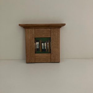 May include: A rectangular wooden wall shelf featuring a central inset tile. The tile displays a graphic illustration of a forest scene with brown tree trunks, green foliage, and light blue sky elements. The frame is constructed from light brown wood.
