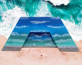 Può includere: Un lavabo trapezoidale caratterizzato da un design in resina che imita una costa oceanica. La superficie passa da un'area di mare profondo blu scuro ad acque basse turchesi, terminando con un bordo di spiaggia sabbiosa con schiuma bianca.