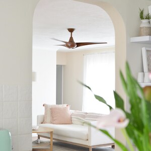 Puede incluir: Un ventilador de techo de madera oscura con tres aspas curvas est&aacute; montado en una habitaci&oacute;n que cuenta con un sof&aacute; blanco con cojines de color rosa claro y rayas, una mesa de centro redonda de madera y un arco blanco que enmarca la vista.