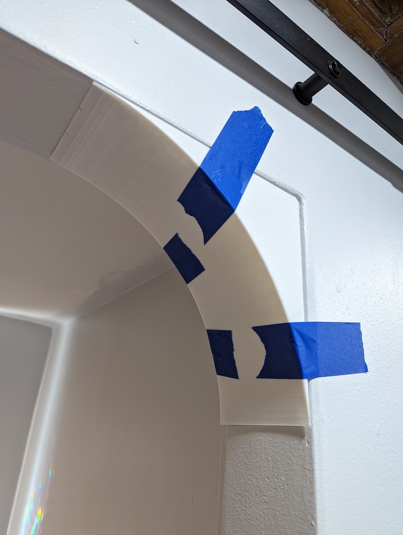 Puede incluir: Un inserto de arco blanco, curvo e impreso en 3D, est&aacute; asegurado temporalmente en la esquina de una abertura de pared blanca con tres tiras de cinta de pintor azul brillante. La pieza tiene un acabado mate liso y sigue el contorno del muro.