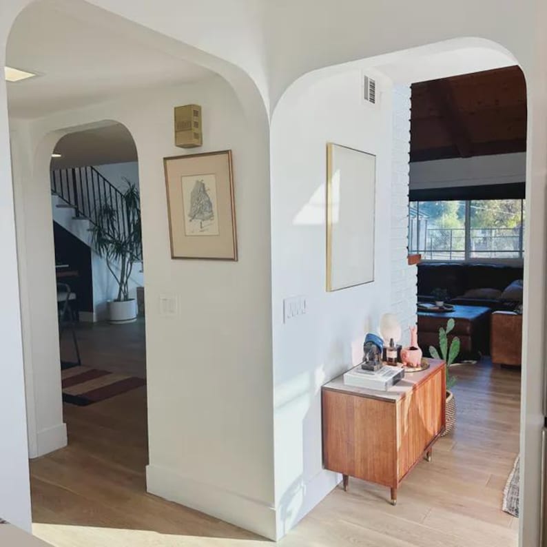Puede incluir: Una vista interior de una casa con arcos blancos, suelos de madera clara y un mueble de madera. Las paredes est&aacute;n decoradas con cuadros enmarcados y el espacio se abre a una sala de estar con un sof&aacute; de cuero oscuro y una ventana grande.