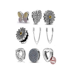 Puede incluir: Un conjunto de nueve clips de pulsera de plata con varios diseños, incluyendo una mariposa, una flor, un corazón y un círculo. Los abalorios están adornados con cristales de colores.