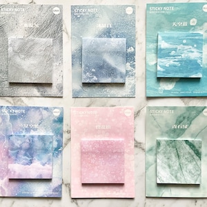 May include: Six square sticky note pads with different designs. The pads feature a grey stone texture, a blue and grey watercolour effect, a blue sky and cloud design, a violet and blue watercolour design, a pink floral pattern, and a green marble pattern.