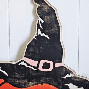 May include: Wooden Halloween decoration of a pumpkin wearing a witch's hat. The hat is black with a pink buckle, and the pumpkin is orange. The decoration is made of wood and has a rustic, handmade appearance.