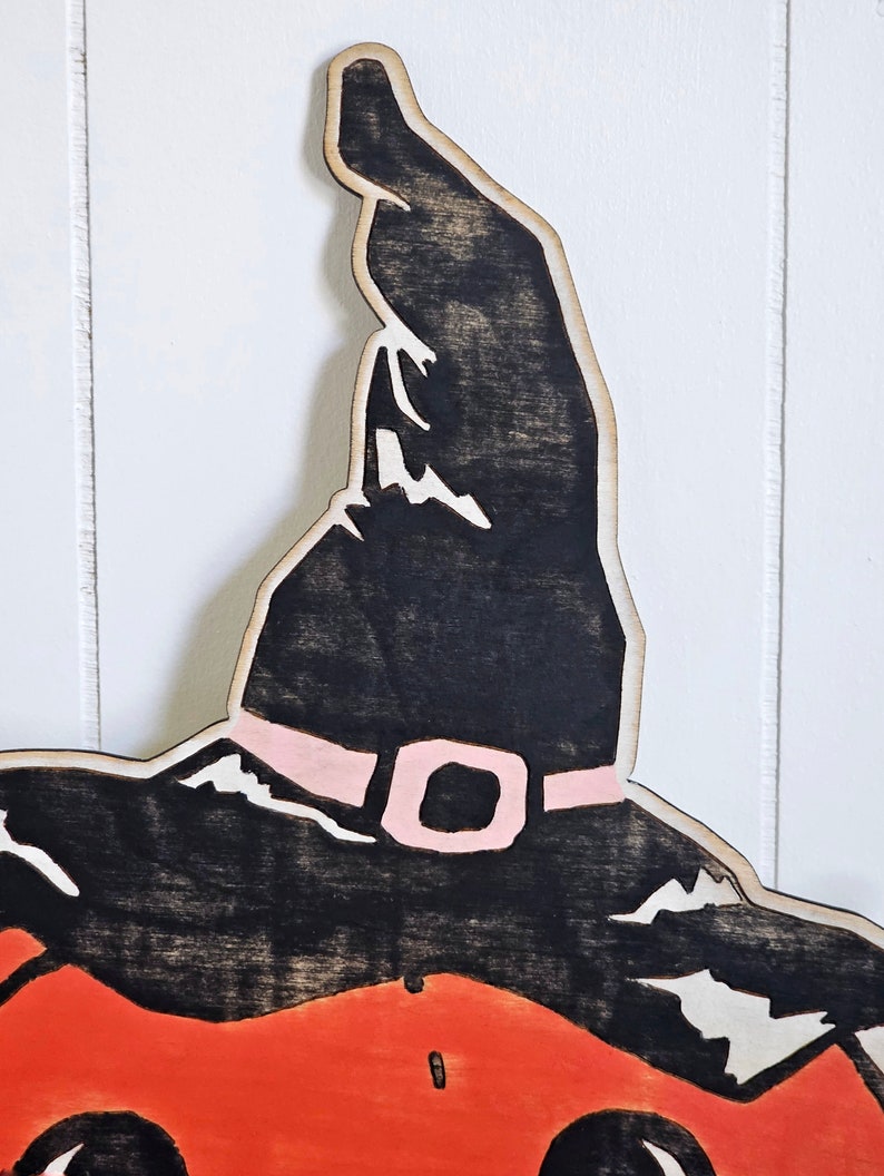 May include: Wooden Halloween decoration of a pumpkin wearing a witch's hat. The hat is black with a pink buckle, and the pumpkin is orange. The decoration is made of wood and has a rustic, handmade appearance.