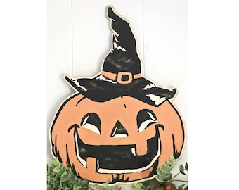 May include: A wooden Halloween decoration featuring a smiling jack-o'-lantern wearing a black witch's hat with an orange buckle. The pumpkin is orange with black facial features and a white outline. The decoration is a festive addition to any Halloween display.