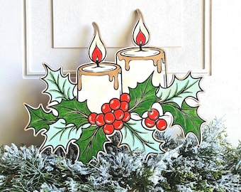 May include: A flat wooden cutout decoration featuring two white candles with orange flames, surrounded by green holly leaves and bright red berries. The design is illustrated with bold black outlines and sits atop a bed of frosted pine branches.