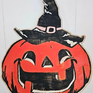 May include: Wooden Halloween decoration of a smiling jack-o'-lantern wearing a black witch's hat with a pink band. The pumpkin is orange with black facial features and a light brown border.