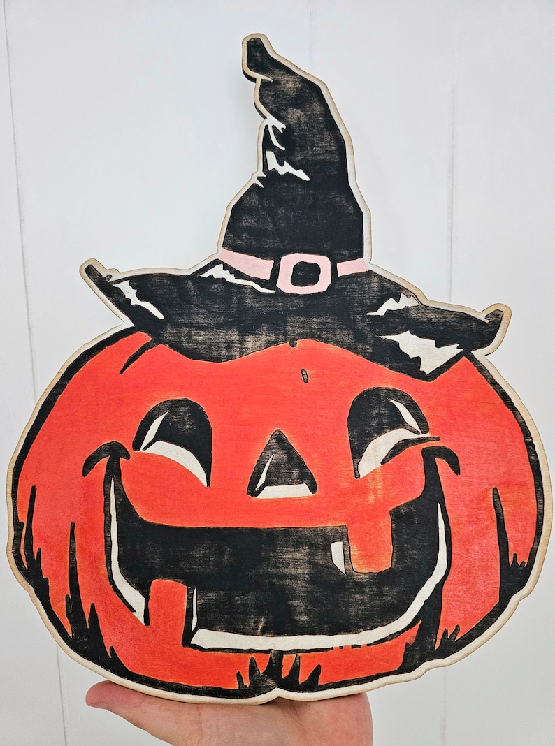 May include: Wooden Halloween decoration of a smiling jack-o'-lantern wearing a black witch's hat with a pink band. The pumpkin is orange with black facial features and a light brown border.