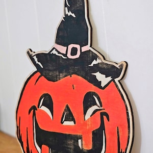 May include: Wooden Halloween decoration of a smiling jack-o'-lantern wearing a witch's hat. The pumpkin is orange with black details, and the hat is black with a pink buckle. A festive, seasonal item.
