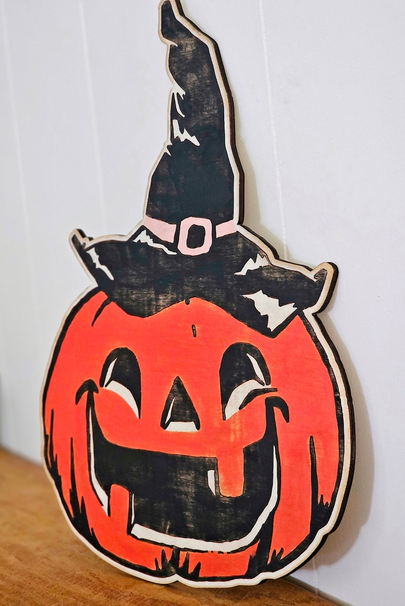 May include: Wooden Halloween decoration of a smiling jack-o'-lantern wearing a witch's hat. The pumpkin is orange with black details, and the hat is black with a pink buckle. A festive, seasonal item.