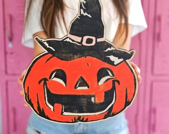 May include: A wooden Halloween pumpkin cutout with a black witch hat and a wide, toothy grin. The pumpkin is painted orange with black details and a pink band around the hat.