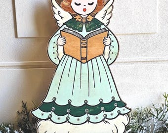 May include: A flat wooden cutout of an angel with auburn hair, white wings, and a light green dress with dark green scalloped trim. The angel has closed eyes, red lips, and holds an open tan book. The figure stands on a white cloud base.