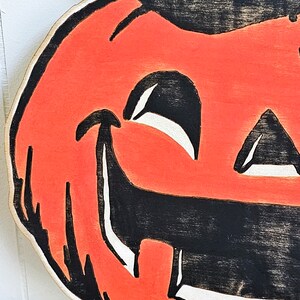 May include: A wooden Halloween decoration featuring a smiling jack-o'-lantern face. The pumpkin has an orange face with black eyes, nose, and mouth. The design is painted with a distressed, vintage look, and the edges are white.