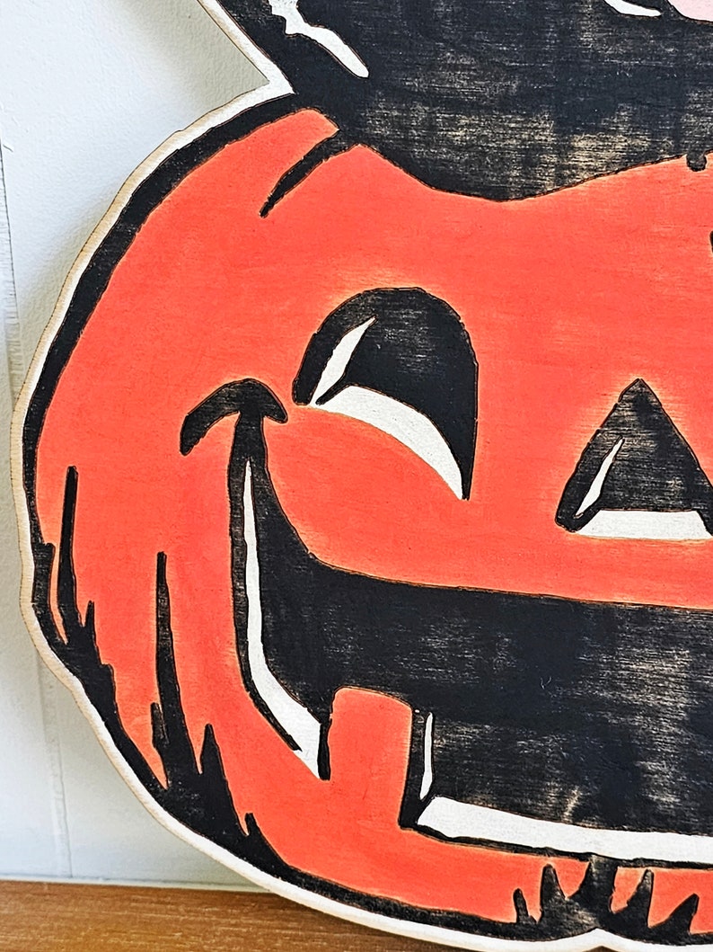 May include: A wooden Halloween decoration featuring a smiling jack-o'-lantern face. The pumpkin has an orange face with black eyes, nose, and mouth. The design is painted with a distressed, vintage look, and the edges are white.