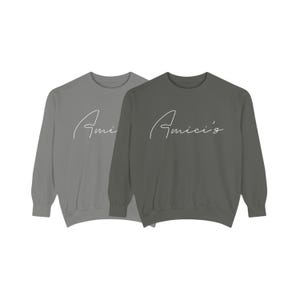 May include: Two long-sleeved sweatshirts, one light gray and one dark gray, are displayed. Each sweatshirt has the word "Amici's" in a white script font across the chest. The sweatshirts are a simple, classic design.