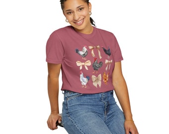May include: A woman wearing a maroon t-shirt with a pattern of chickens and bows. The shirt has a crew neck and short sleeves.