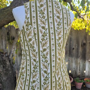 Puede incluir: Un chaleco vintage con un estampado floral en tonos verdes y beige sobre un fondo blanco. El chaleco sin mangas tiene una silueta entallada. La tela presenta rayas verticales con motivos florales y de hojas.