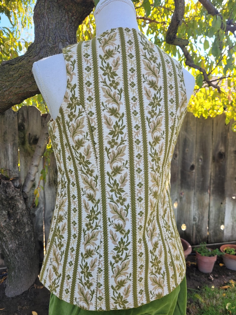 Puede incluir: Un chaleco vintage con un estampado floral en tonos verdes y beige sobre un fondo blanco. El chaleco sin mangas tiene una silueta entallada. La tela presenta rayas verticales con motivos florales y de hojas.
