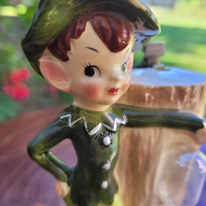May include: A ceramic figurine of a young boy in a green outfit and hat, with a whimsical design. The boy has rosy cheeks, brown hair, and is posed with one hand on his hip. The figurine is set against a blurred outdoor background.