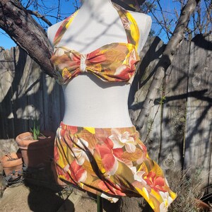 May include: A vintage two-piece swimsuit with a floral pattern in shades of yellow, orange, and brown. The halter top has a bow detail, and the skirt is gathered at the waist. The swimsuit is displayed on a mannequin outdoors.