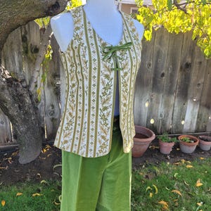 Puede incluir: Un conjunto vintage de dos piezas con un chaleco estampado y pantalones de pernera ancha. El chaleco tiene un fondo crema con rayas florales verdes y doradas y un cierre de cinta verde. Los pantalones son de terciopelo verde brillante liso.