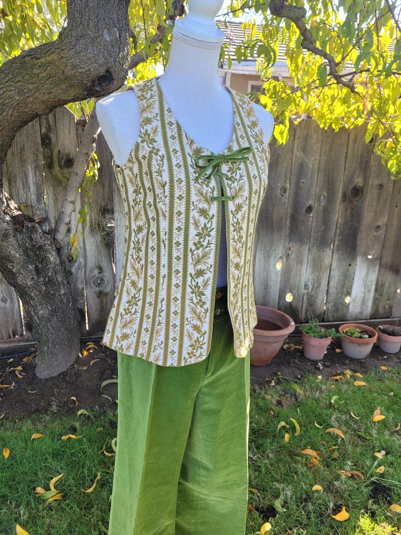 Puede incluir: Un conjunto vintage de dos piezas con un chaleco estampado y pantalones de pernera ancha. El chaleco tiene un fondo crema con rayas florales verdes y doradas y un cierre de cinta verde. Los pantalones son de terciopelo verde brillante liso.
