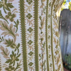 Puede incluir: Primer plano de un vestido vintage con un estampado floral texturizado. El vestido presenta rayas verticales en verde oliva y beige, con dise&ntilde;os florales y geom&eacute;tricos. La tela parece ser un material tejido.