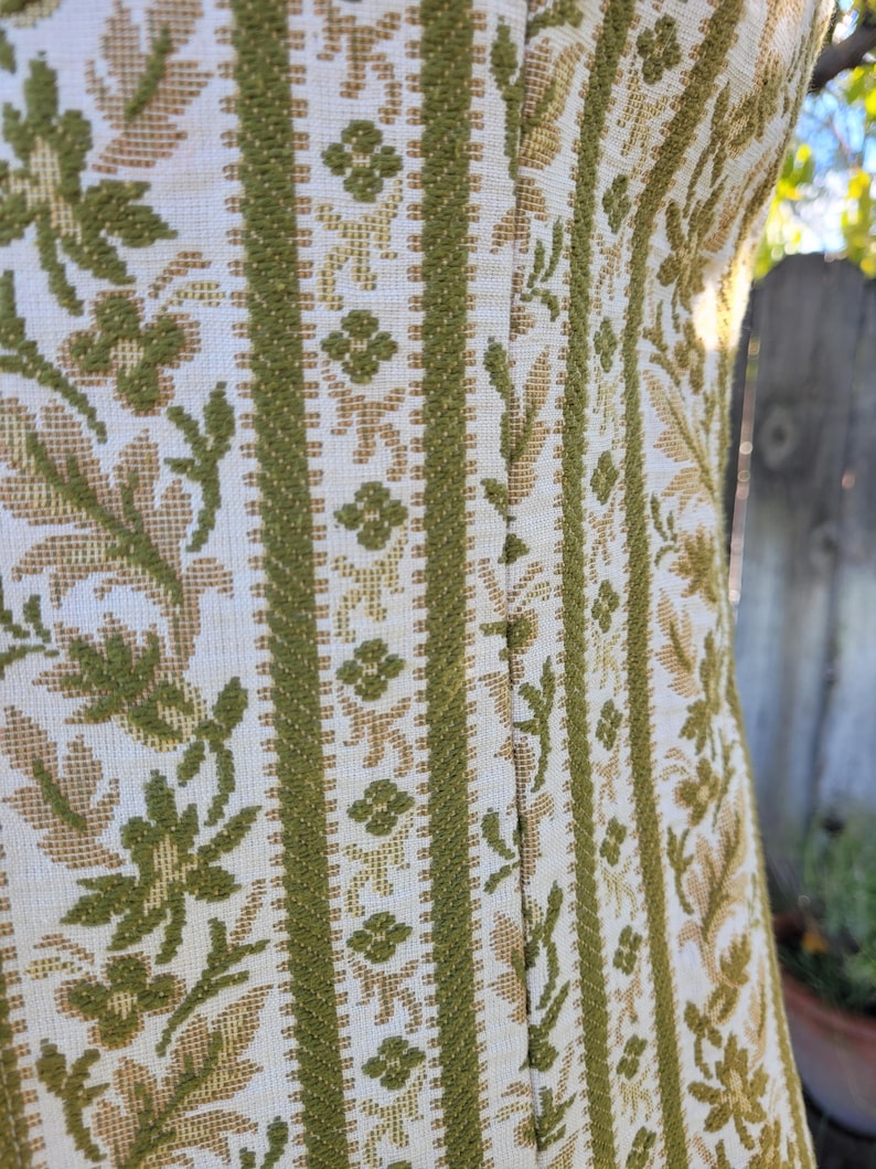 Puede incluir: Primer plano de un vestido vintage con un estampado floral texturizado. El vestido presenta rayas verticales en verde oliva y beige, con dise&ntilde;os florales y geom&eacute;tricos. La tela parece ser un material tejido.