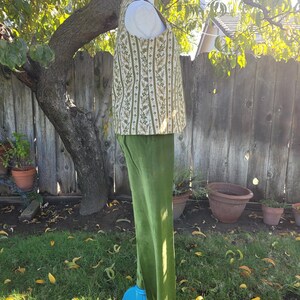 Puede incluir: Un conjunto de dos piezas con un chaleco crema con un estampado floral vertical en verde y dorado, combinado con pantalones de terciopelo verde oliva. El conjunto se exhibe en un maniqu&iacute; azul en un entorno al aire libre.