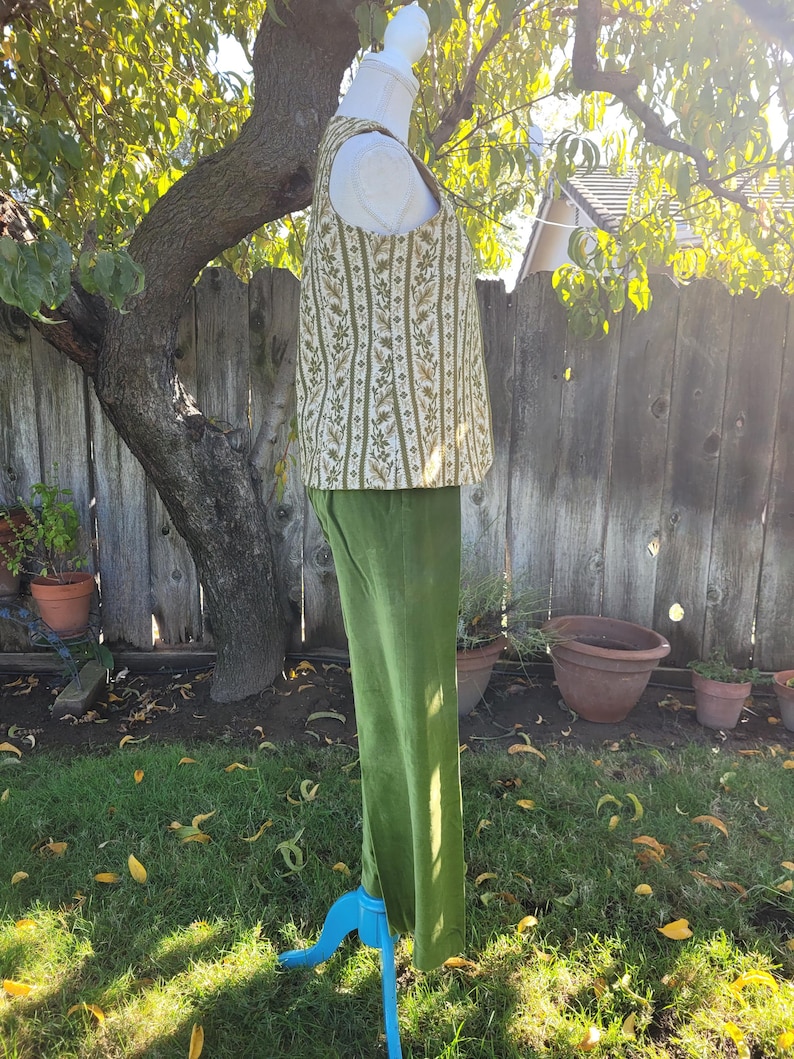 Puede incluir: Un conjunto de dos piezas con un chaleco crema con un estampado floral vertical en verde y dorado, combinado con pantalones de terciopelo verde oliva. El conjunto se exhibe en un maniqu&iacute; azul en un entorno al aire libre.
