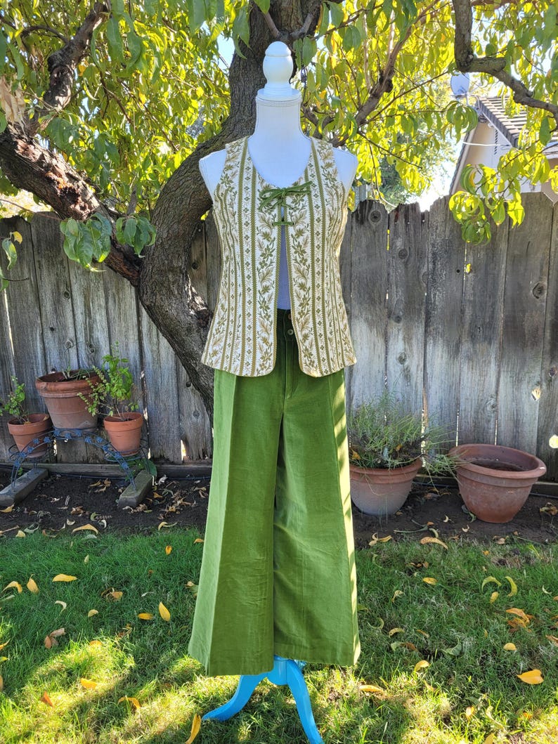 Puede incluir: Un atuendo vintage con un chaleco color crema con bordado floral verde y un par de pantalones de pana verdes de pierna ancha a juego. El chaleco tiene un cierre de lazo. El atuendo se exhibe en un maniqu&iacute; al aire libre.