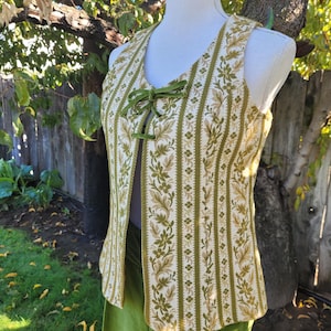 Puede incluir: Un chaleco vintage con un estampado floral en tonos verdes y beige. El chaleco tiene un dise&ntilde;o de rayas verticales y un cierre de lazo de terciopelo verde. Se muestra en un maniqu&iacute;, combinado con pantalones verdes.