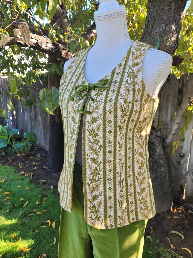 Puede incluir: Un chaleco vintage con un estampado floral en tonos verdes y beige. El chaleco tiene un dise&ntilde;o de rayas verticales y un cierre de lazo de terciopelo verde. Se muestra en un maniqu&iacute;, combinado con pantalones verdes.