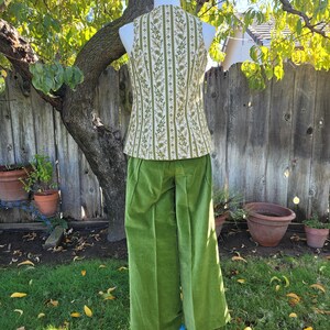 Puede incluir: Un atuendo vintage en un maniqu&iacute;, con un chaleco con estampado floral verde y blanco y pantalones verdes de pernera ancha. El chaleco tiene un patr&oacute;n de rayas verticales. Los pantalones son de color verde liso y vibrante. El atuendo se exhibe al aire libre sobre un soporte azul.