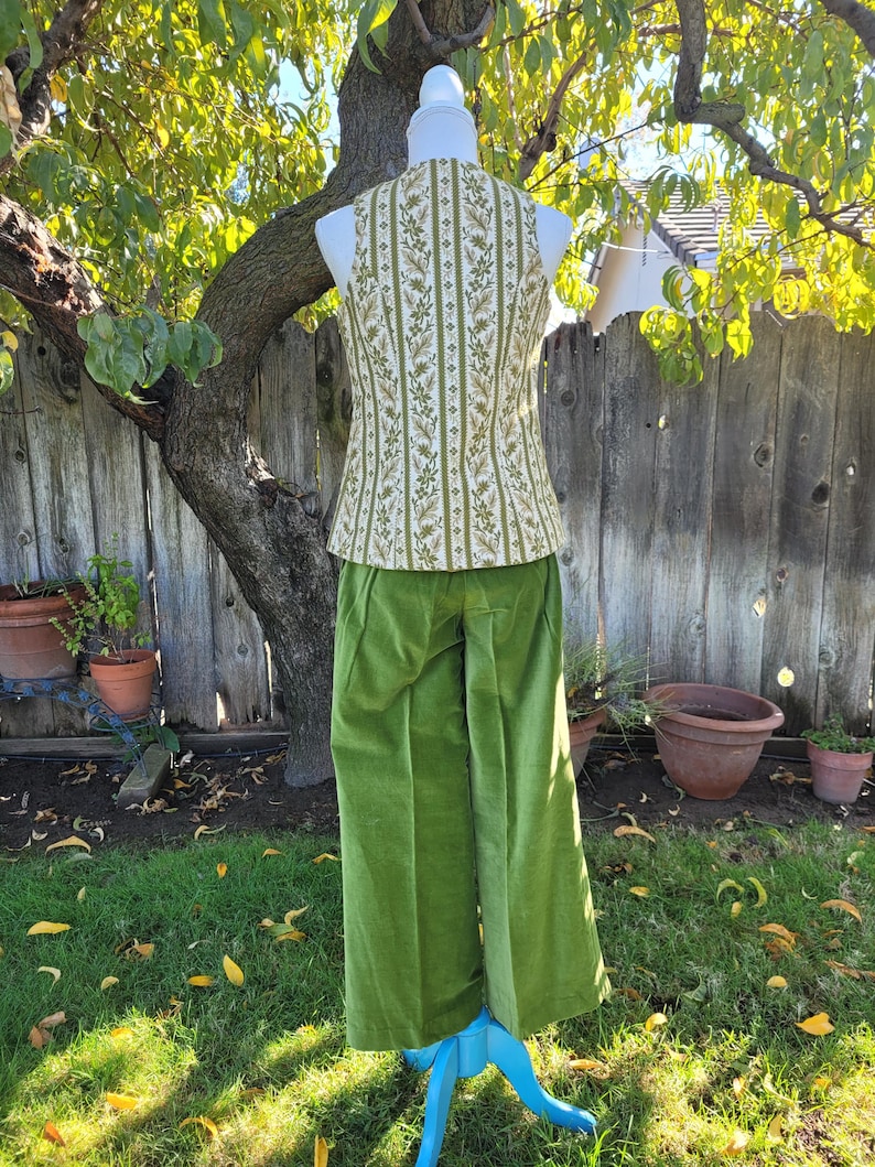 Puede incluir: Un atuendo vintage en un maniqu&iacute;, con un chaleco con estampado floral verde y blanco y pantalones verdes de pernera ancha. El chaleco tiene un patr&oacute;n de rayas verticales. Los pantalones son de color verde liso y vibrante. El atuendo se exhibe al aire libre sobre un soporte azul.
