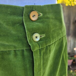 Puede incluir: Primer plano de un pantal&oacute;n de terciopelo verde con dos botones color beige. La tela tiene una textura suave y afelpada. Los botones redondos tienen un brillo sutil. El pantal&oacute;n es de un tono verde vibrante.