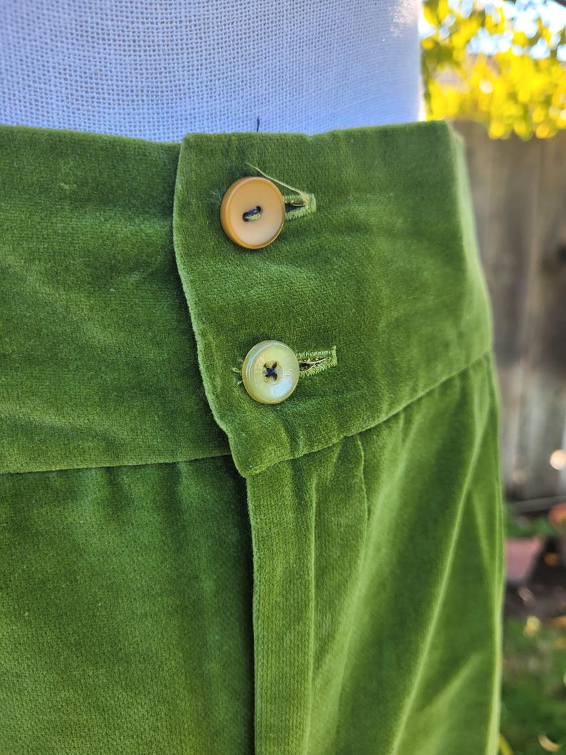 Puede incluir: Primer plano de un pantal&oacute;n de terciopelo verde con dos botones color beige. La tela tiene una textura suave y afelpada. Los botones redondos tienen un brillo sutil. El pantal&oacute;n es de un tono verde vibrante.