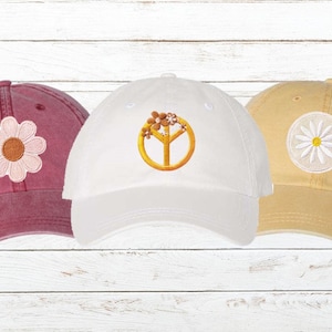 May include: Three baseball caps in burgundy, white, and tan. Each cap features an embroidered design: a daisy, a peace sign with flowers, and a daisy. The caps are displayed against a white wood background.