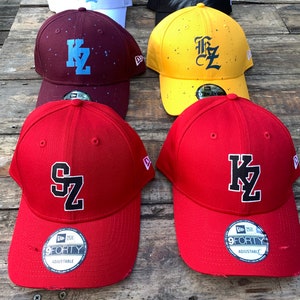 May include: Five baseball caps in different colours and designs. The caps are all adjustable and have a logo on the front. The logos include the letters "SZ", "KZ", and "FZ".