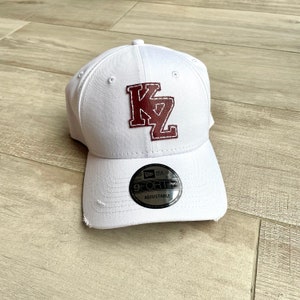 Może przedstawiać: Biała czapka bejsbolówka z naszywką ze skóry bordowej, na której widnieje napis "KZ" w haftowanym foncie. Czapka ma regulowany pasek w kolorze czarnym i srebrnym z logo New Era i napisem "9FORTY ADJUSTABLE".