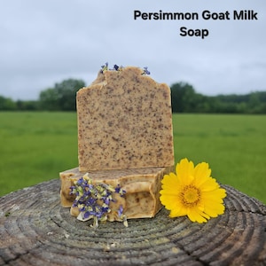 May include: A rectangular bar of tan-coloured Persimmon Goat Milk Soap with dark brown speckles throughout. The soap is topped with small blue dried flower petals and is positioned next to a single bright yellow flower blossom.