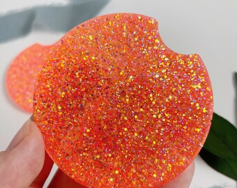 May include: A round orange glitter coaster with a cut-out for a cup. The coaster is made of resin and has a sparkly finish.